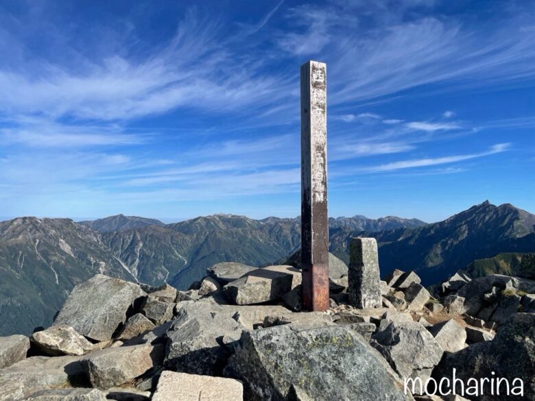 西穂高岳山頂から見た眺望と看板の写真。青空と広がる山並みの様子。