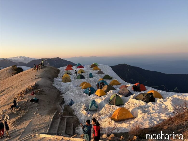 夕焼けに染まるテントサイトの写真。残雪の上に張られた色とりどりのテントが並んでいる様子。